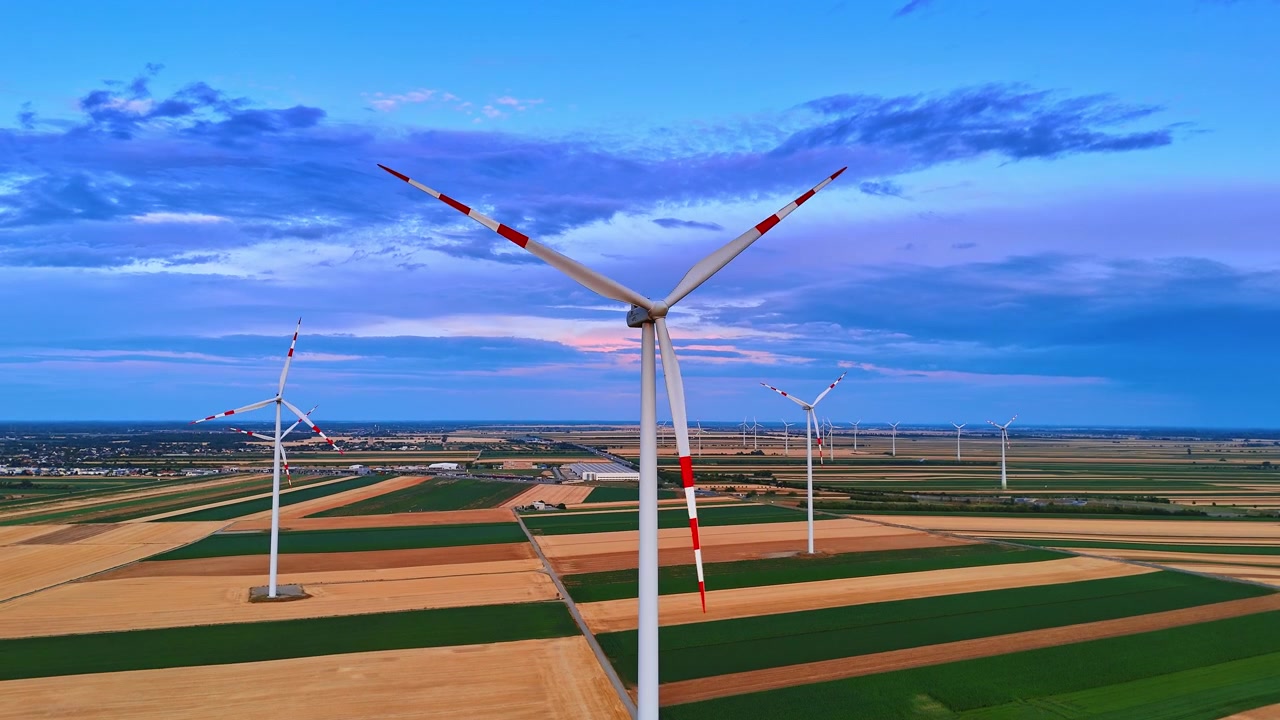 Parco eolico a energia pulita su terreno agricolo a strisce. Prospettiva aerea di turbine ecologiche su campi rettangolari verdi e marroni sotto un'atmosfera serale blu.