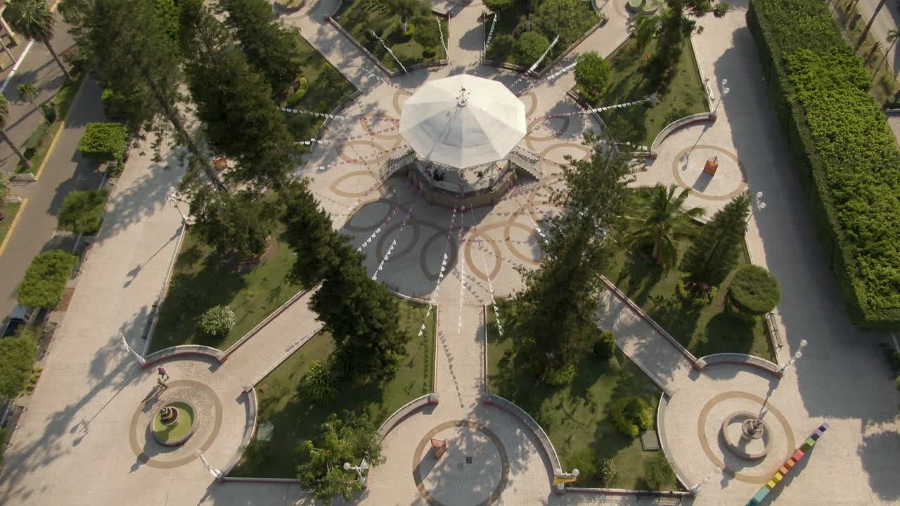 Overhead aerial tilt reveals symmetrical design of central park with gazebo, pathways, and pine trees in Tuxpan, Jalisco