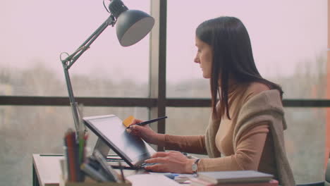 a professional designer in the office draws with a stylus on a graphic tablet sitting in an office with huge windows in the loft style. modern office of graphic designer and creator.