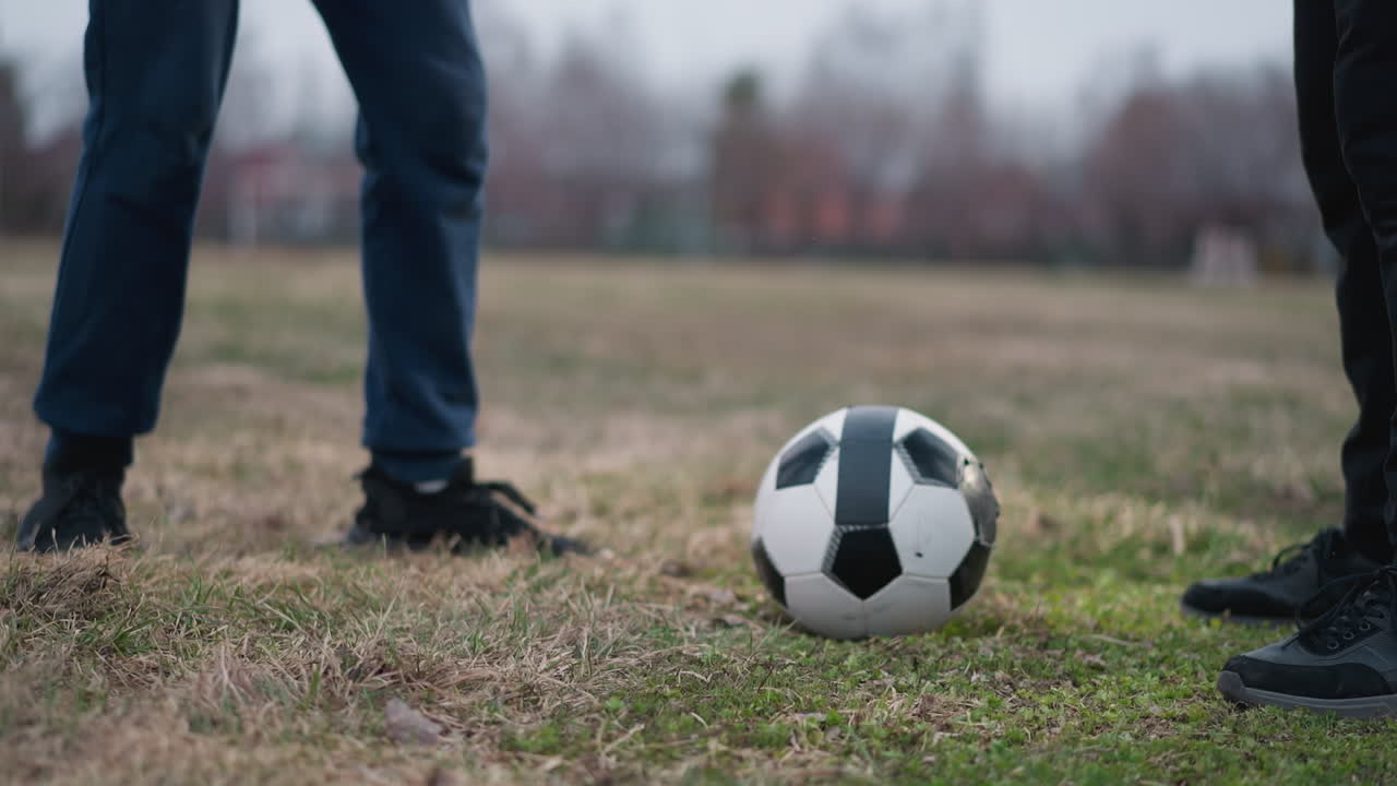 Close-up leg view of two people preparing to play soccer with a ball placed between them on a grassy field, the background is blurred, showcasing a football post in the distance