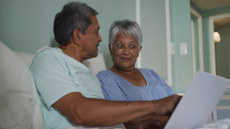 Happy-senior-mixed-race-couple-sitting-in-bed-using-laptop