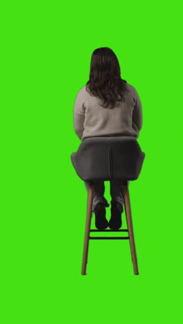 Vertical-Video-Rear-View-Full-Length-Shot-Of-Casually-Dressed-Young-Woman-Sitting-On-Stool-And-Waiting-Against-Green-Screen