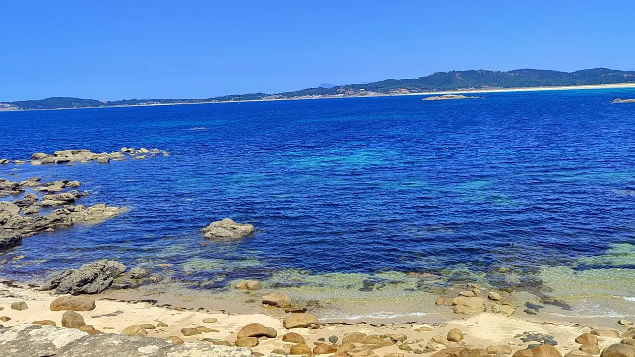 una toma de una playa rocosa con agua clara y oscura, cielo despejado y tierra en el horizonte.