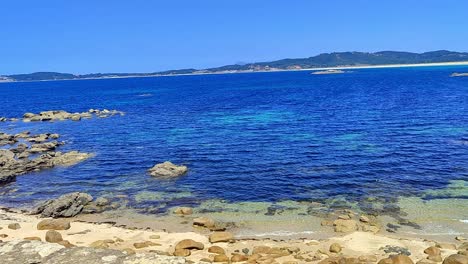 Pan-Shot-of-Rocky-Beach-with-Clear-Dark-Water-Clear-Sky-and-Land-in-Horizon