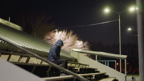 a man wearing a hooded jacket sits alone on stadium bleachers at night, moving slightly while illuminated by streetlights, the background shows a dimly lit building, particles floating