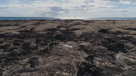 drone flying low over volcanic rocky shore, towards pacific ocean, sunlight reflection on water