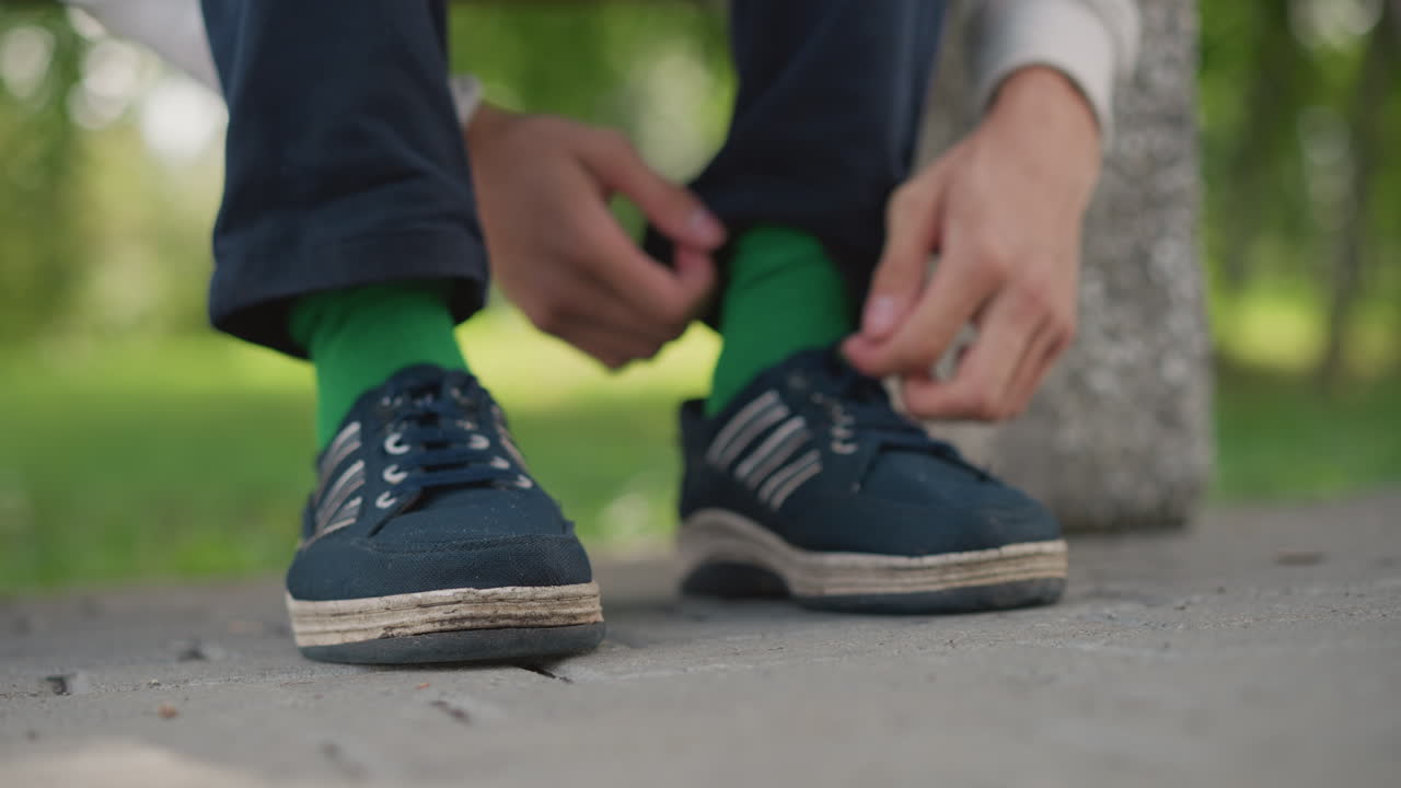 Hombre sentado en un banco atándose el cordón del zapato; primer plano de los dedos tirando de los cordones, calcetines verdes y zapatillas deportivas azul marino, pavimento y árboles del parque al fondo, luz nublada, corredor concentrado preparándose para trotar, rápido