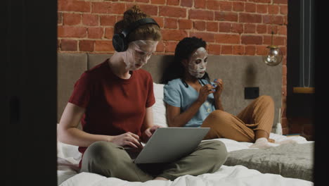 y chicas con máscara facial escuchando música en una laptop y limando sus uñas en la cama
