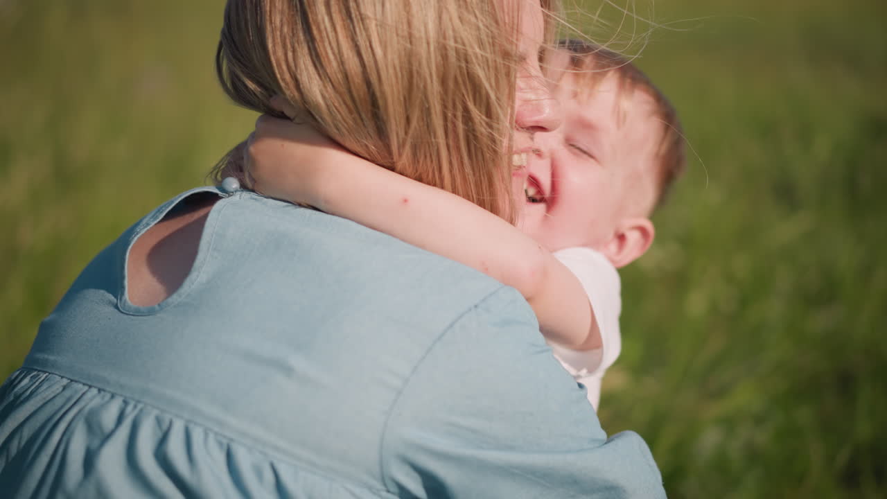 A woman in a blue dress bends down to embrace her son,who hugs her blissfully with a big smile on his face. tender moment of love and connection between mother and child in a sunny, green grass field
