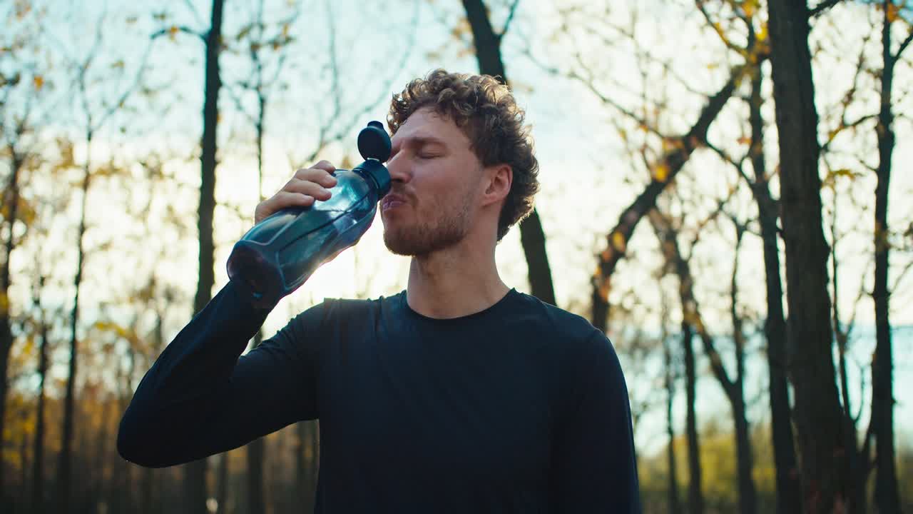un hombre feliz con cabello rizado y barba en un uniforme deportivo negro bebe agua de una botella deportiva gris y se vierte sobre ella para relajarse después de correr por la mañana en otoño