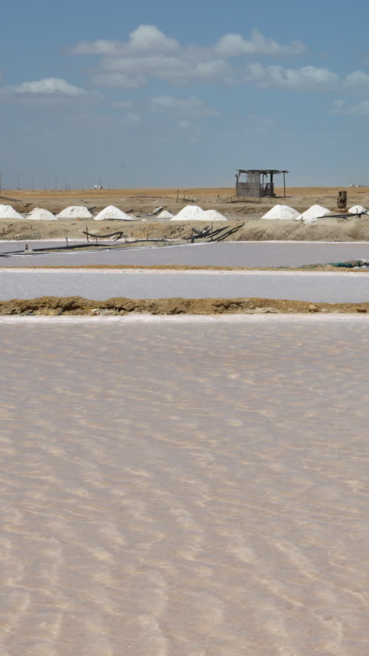 Vídeo vertical deslumbrante de salinas e pilhas de sal brancas em Manaure, La Guajira, Colômbia. Luz solar intensa, céu azul, cabana rústica e água cintilante criam uma cena hipnotizante pronta para redes sociais.