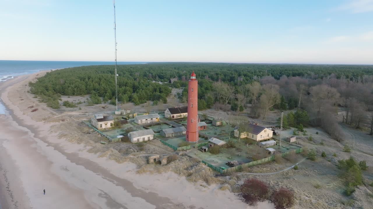 vista aérea del faro akmenrags de color rojo, costa del mar báltico, letonia, playa de arena blanca, mar en calma, día soleado con nubes, disparo en órbita de drones distantes