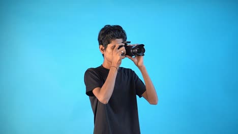 Un-Joven-Indio-Divertido-Con-Una-Camiseta-Negra-Básica-Aislado-En-Un-Estudio-De-Fondo-Azul-Y-Tomando-Fotos-Con-Su-Dslr