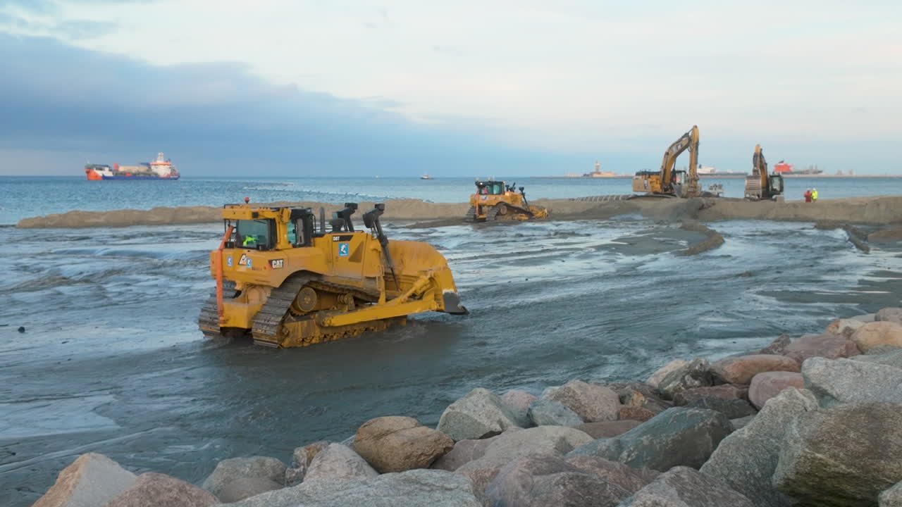 A large yellow bulldozer and other heavy machinery actively work on coastal reclamation, pushing mud and sand near a rocky breakwater at twilight