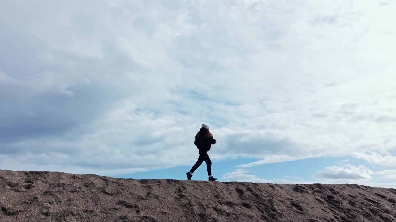 Cheerful woman running on a sandy hill on the seashore. Woman in a jacket and hat walking on the sand by the sea in autumn. Autumn walk by the sea.