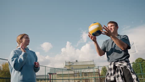 close-up of volleyball training session for beginners with coach wearing plaid shirt around waist, passing ball amongst players in open court with background of building and fence