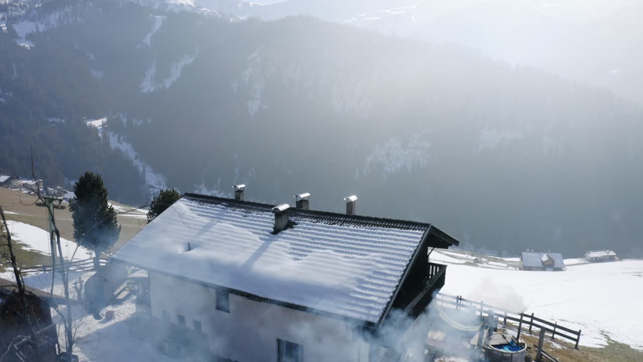 Drone flies over a cozy mountain hut with a steaming hot tub. Mist drifts through the valley, snow lingers on the grass, and white peaks tower above.