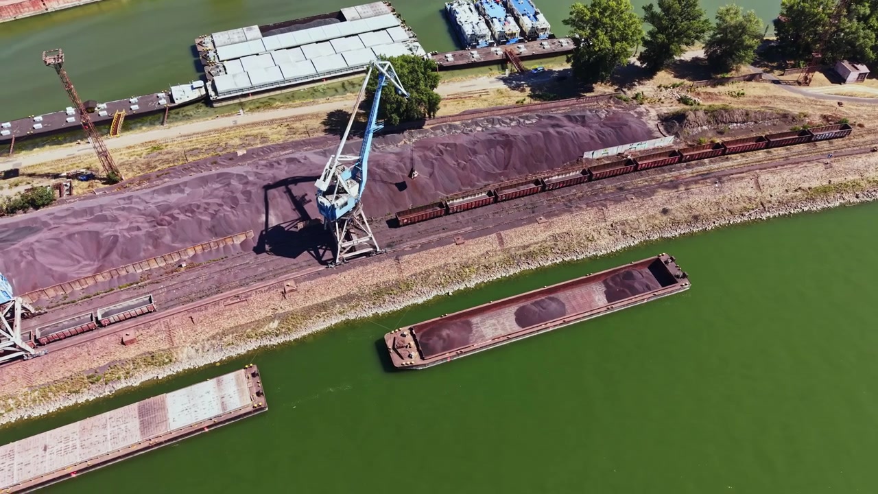 Il carico viene caricato su una chiatta in un cantiere navale vicino a un fiume durante il giorno, con attrezzature e materiali industriali in vista.