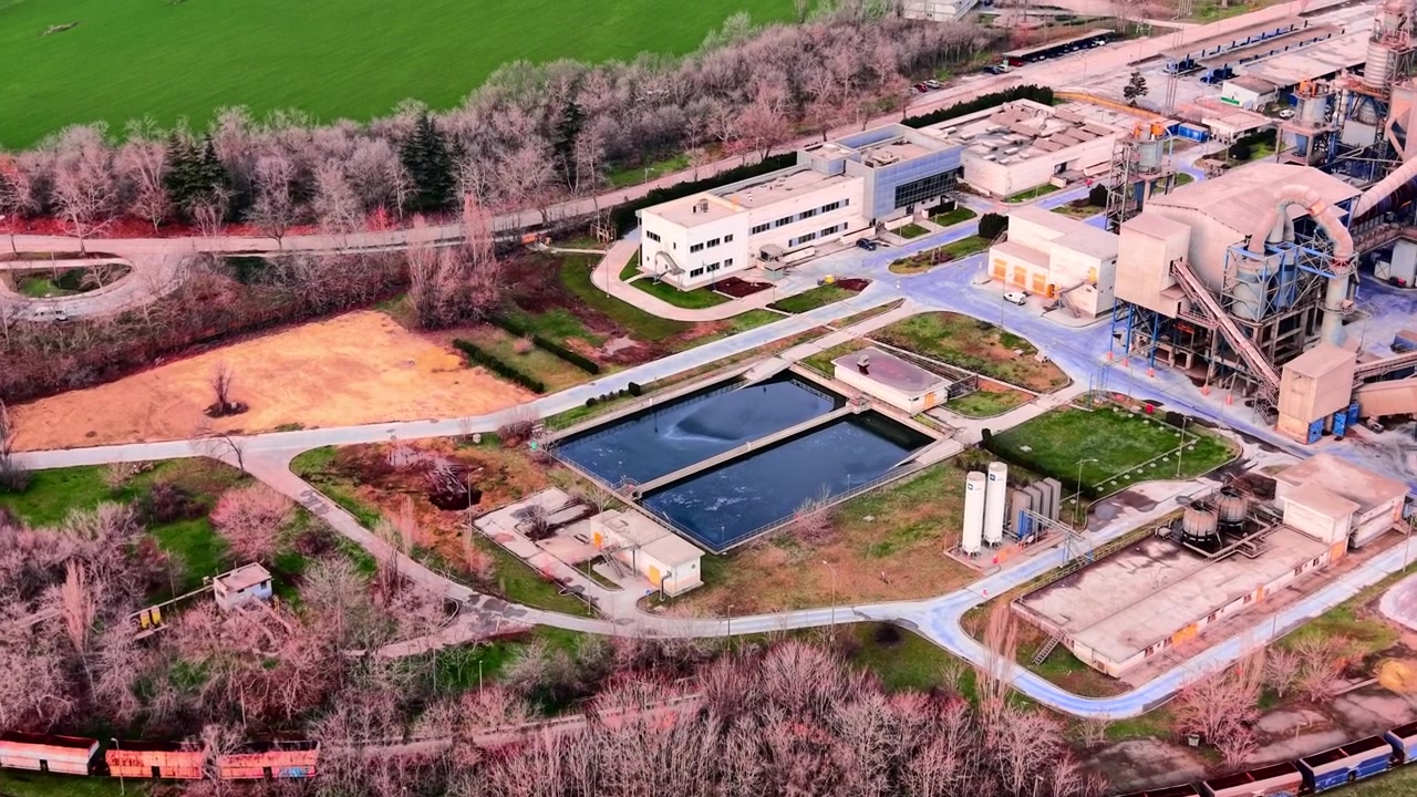 Luftfoto af cementfabrik med siloer og sedimentationsbassin, Bulgarien. Industrianlæg med hvid bygning, cirkulært vandbassin, lagertanke, godsvogne og omgivende grønne træer