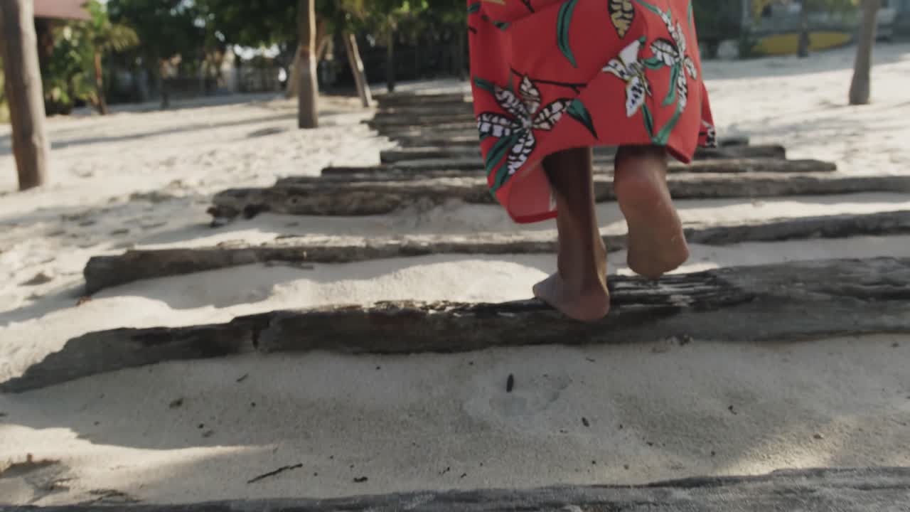 Parte inferiore del corpo di donna ispanica in pareo che cammina a piedi nudi su una passerella su una spiaggia soleggiata, al rallentatore