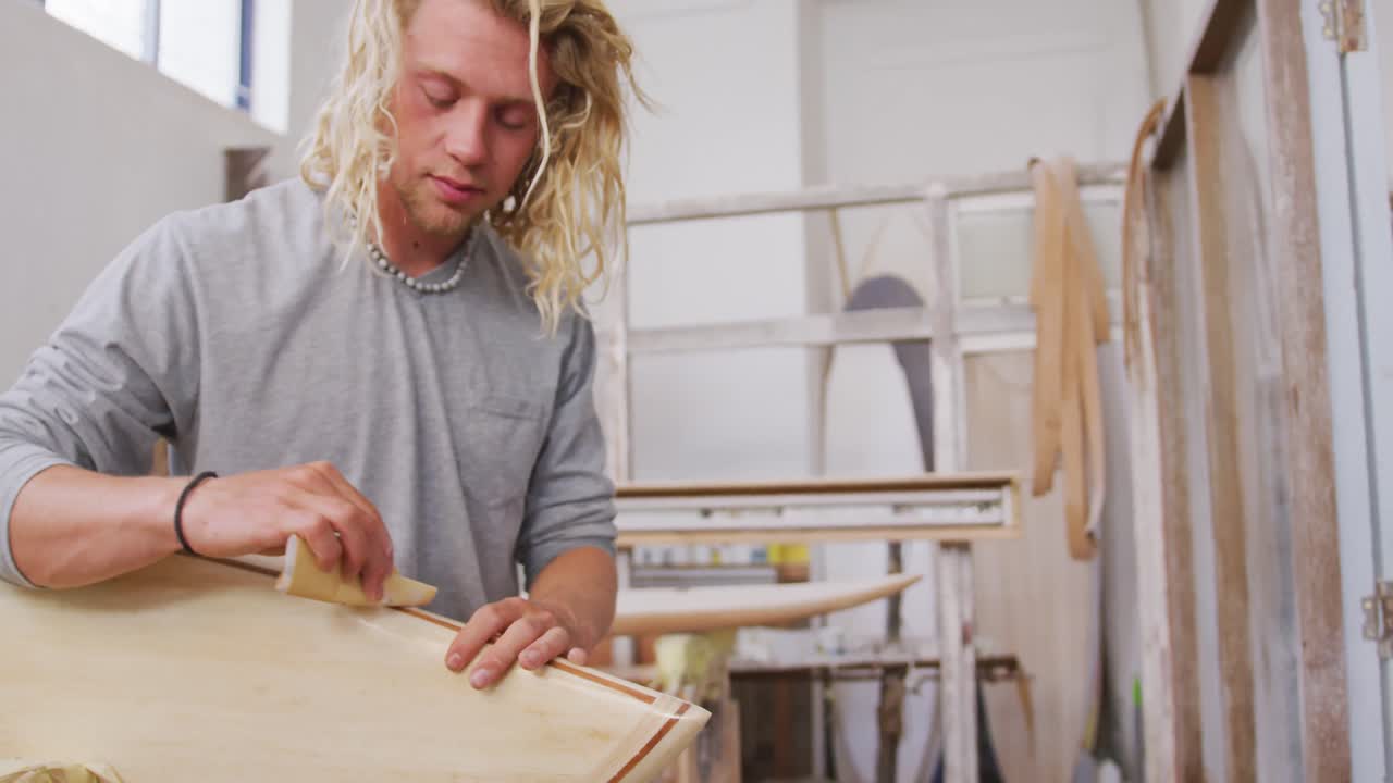 A Caucasian male surfboard maker polishing a wooden surfboard edge
