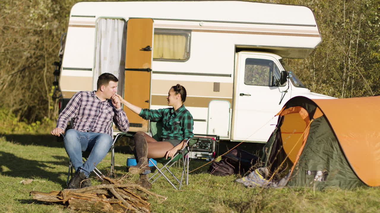 novio hipster besando la mano de su novia mientras está sentado en asientos de camping