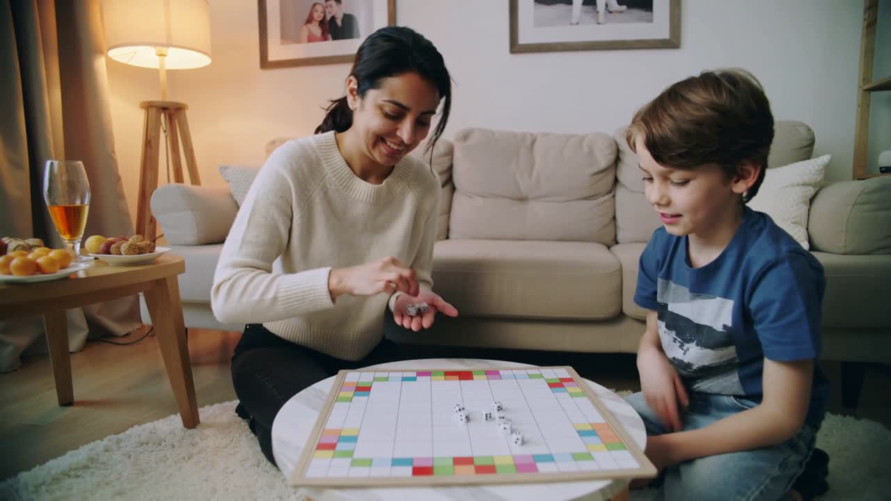 Donna con maglione leggero e figlio con maglietta a righe che allungano le mani per spostare i pezzi di un gioco mentre giocano a casa.