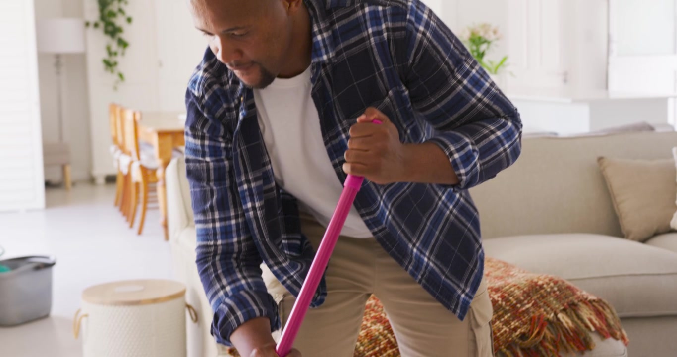 African American man in living room gripping pink mop, mopping tiled floor with blue spray bottle