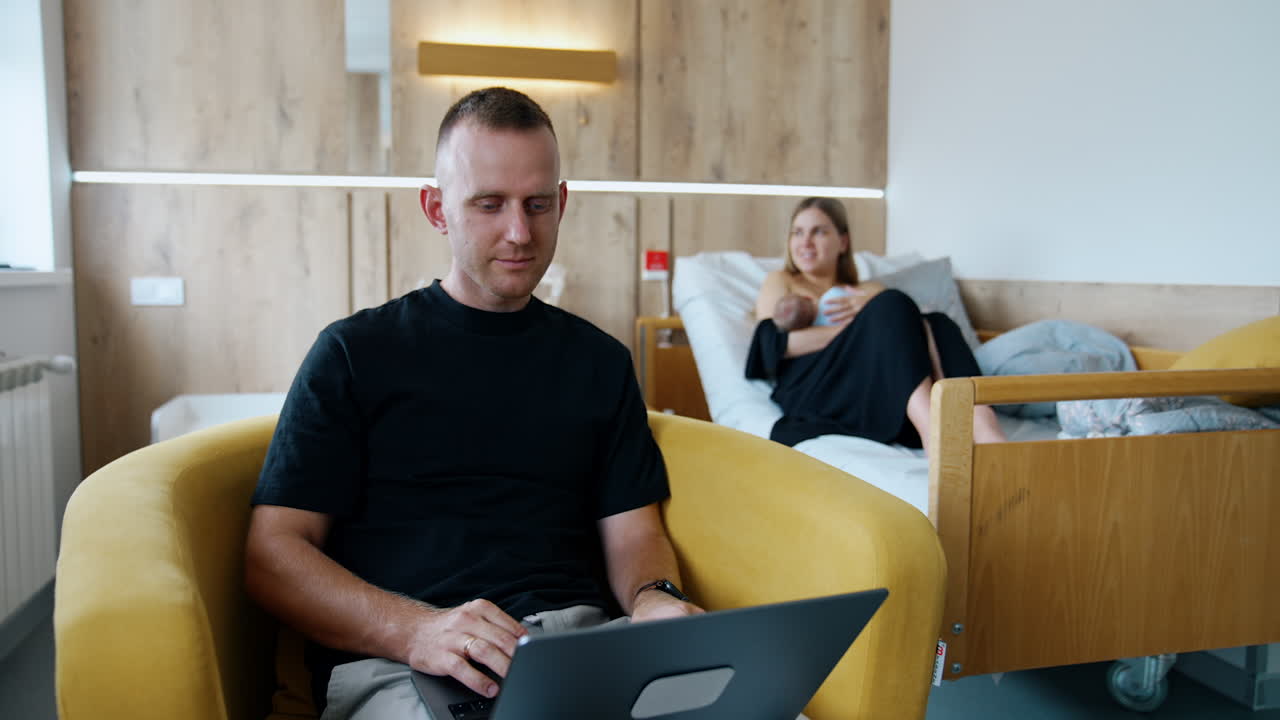 Busy man is focused on his laptop sitting in the armchair. Woman at backdrop feeding her baby lying in bed.