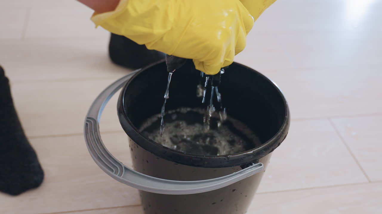 Close up of person wearing yellow gloves squeezing soaked rag inside black bucket with water dripping down on wooden floor, household cleaning process