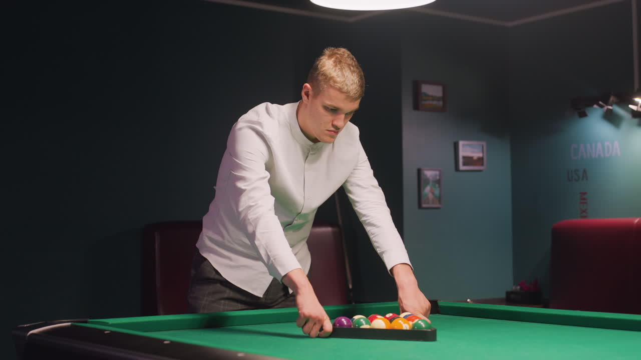 el jugador con camisa blanca y pantalones grises arregla bolas de billar de colores en triángulo en la mesa de billar verde. las luces superiores arrojan un brillo cálido mientras se centra con calma concentración en el elegante salón