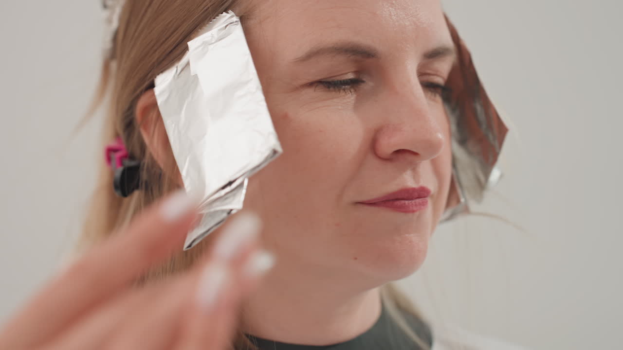 Primo piano su una donna caucasica che fa dei colpi di sole con la stagnola, con la parrucchiera che posiziona fogli di alluminio sulle tempie e sulle sezioni laterali, espressione facciale e mani in primo piano, trattamento dei capelli sotto illuminazione neutra da studio, atmosfera rilassata nel salone