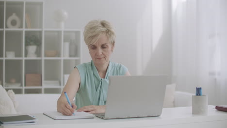Eine-Frau-Mittleren-Alters-Arbeitet-Mit-Einem-Laptop,-Schaut-Sich-Ein-Webinar-An-Und-Schreibt-Notizen-Zur-Entfernten-Selbstbildung