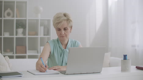 middle-aged woman is working with laptop viewing webinar and writing notes distant self-education