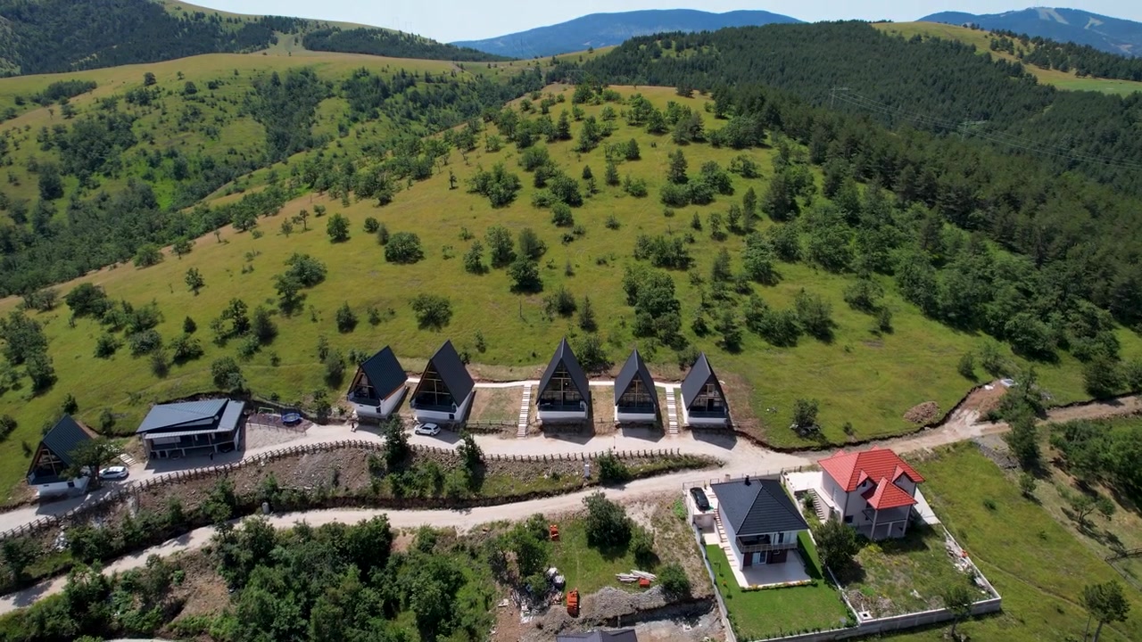 Dron krouží nad kompaktním horským letoviskem s chatami s tmavými střechami, vinoucími se cestami, zelenými loukami a zalesněnými kopci v Zlatiboru, Srbsko, za jasného denního světla.
