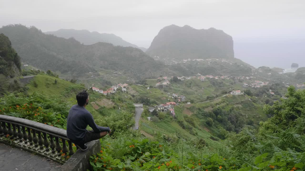 Madeira, Portugal - Man Sitting Alone At The Balcony Overlooking The Beautiful Scenery Of Green Fields And Mountains On A Foggy Day - Closeup Shot