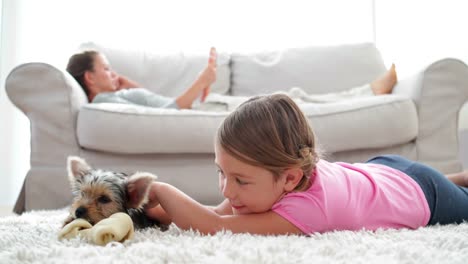 niña jugando con un cachorro masticando hueso con su madre leyendo en el sofá