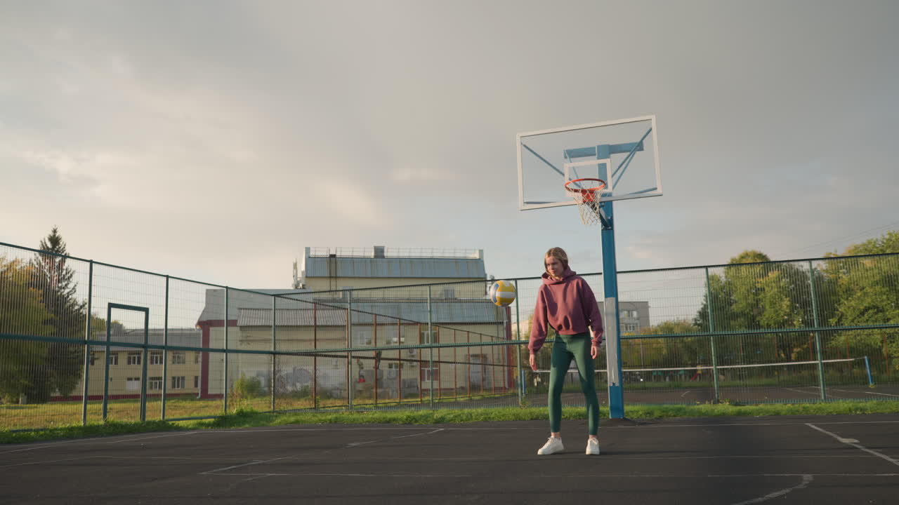 primer plano de una mujer con ropa deportiva casual rebotando una pelota de voleibol al aire libre frente a un edificio, muestra un juego activo en un espacio abierto, con una cancha de voleobol y un cielo despejado