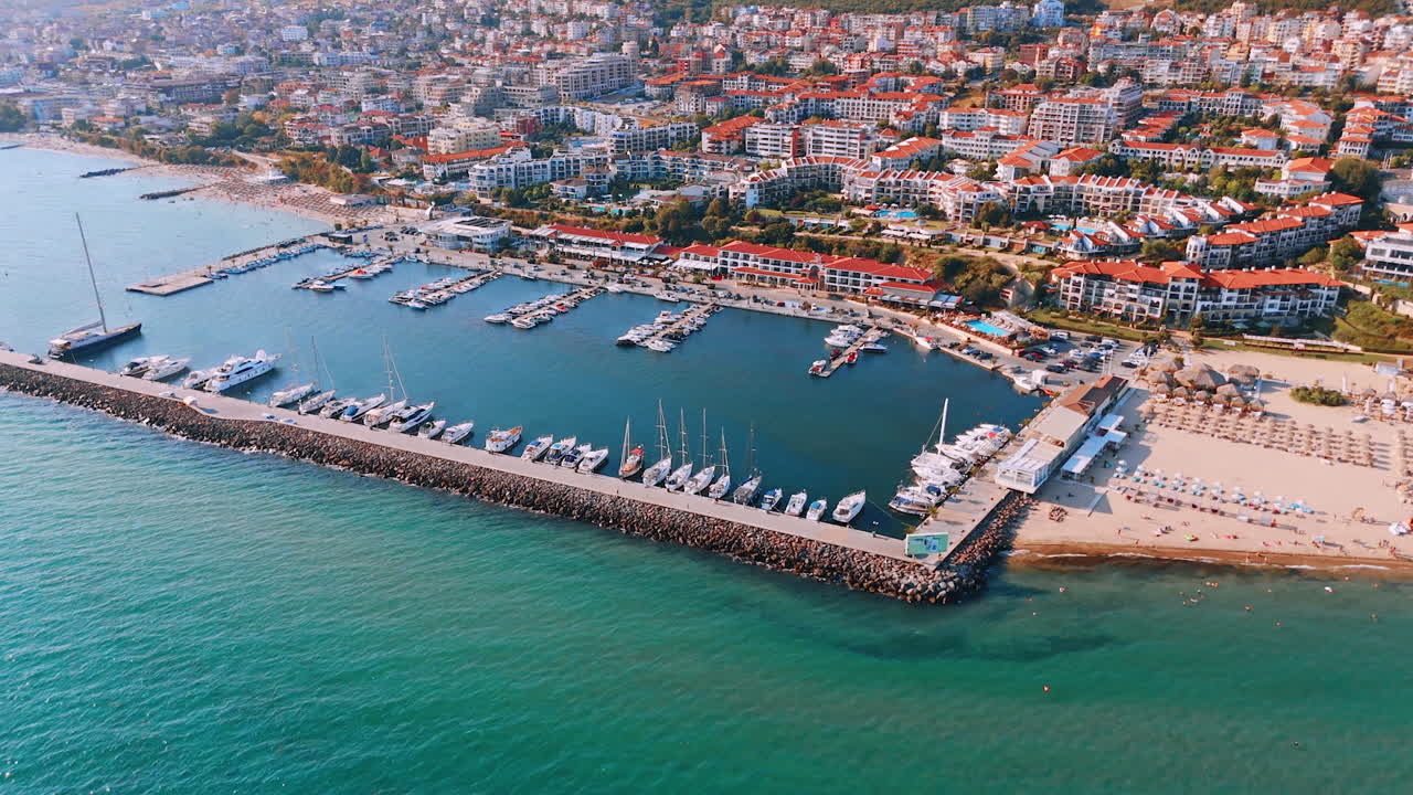 Turquoise water waves hit the stony dam. Boats and yachts stand at the piers. Multiple hotels of sunny Sveti Vlas, Bulgaria at backdrop