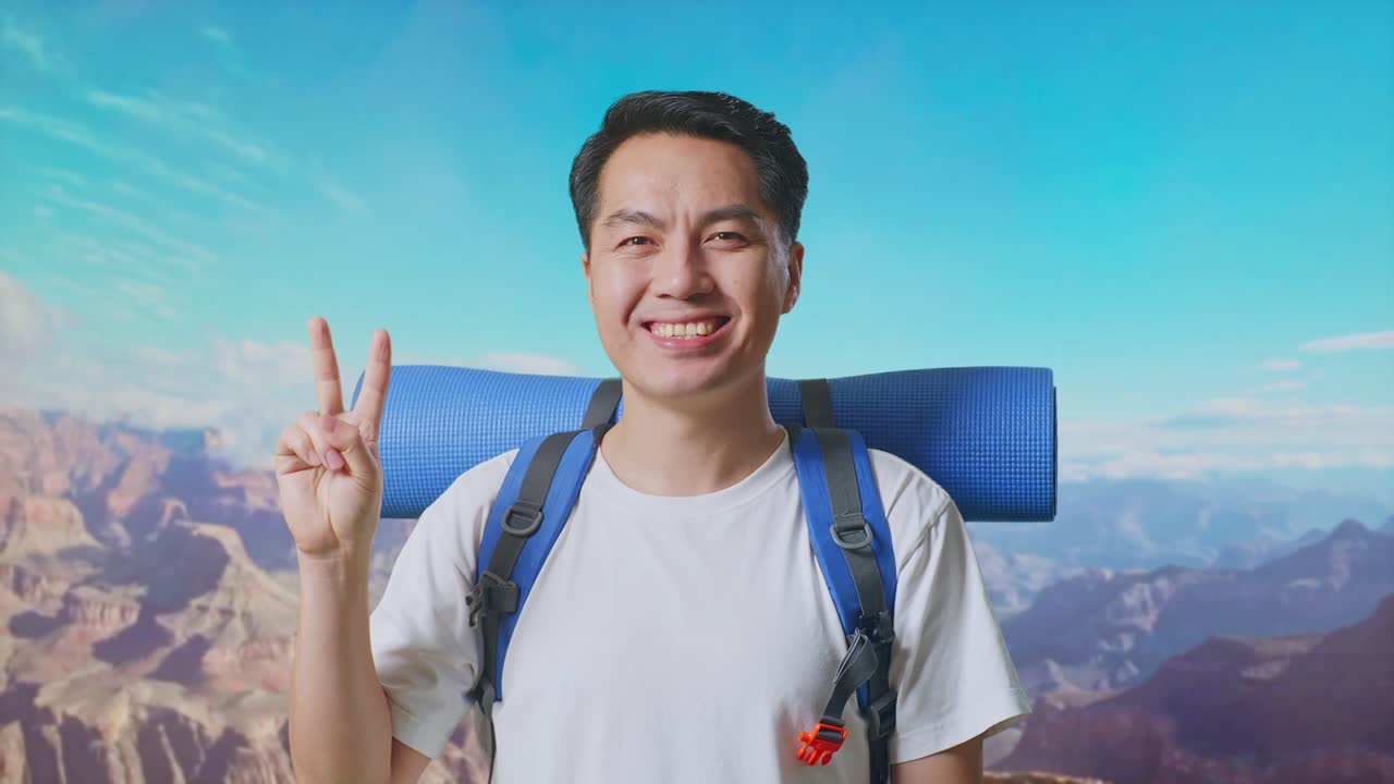 Close Up Of Asian Male Hiker With Mountaineering Backpack Smiling And Showing Peace Gesture While Traveling At The Top Of Mountain