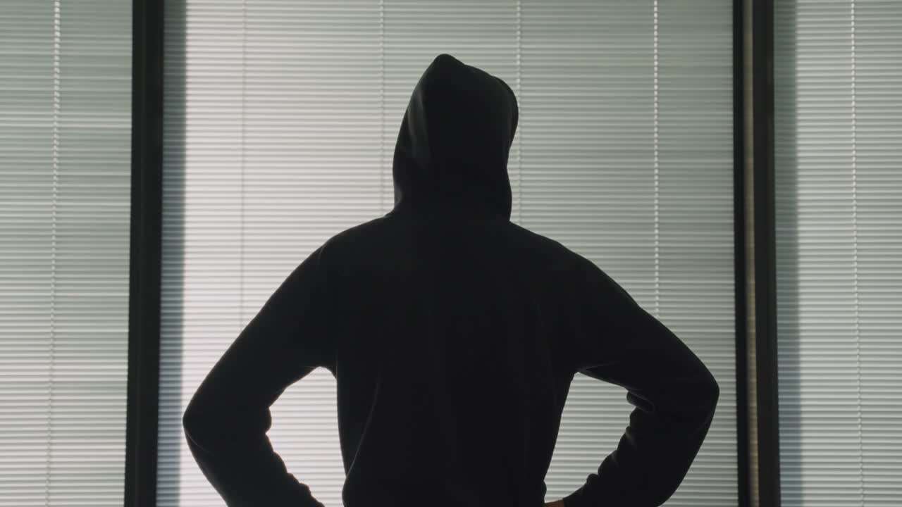 黒いパーカーを着た謎の人物、ハッカーまたはスパイがアパートの窓際に立ち、外を眺めて待っている。犯罪行為や取引。不審な活動を監視している。腕を腰に当てた匿名の男。