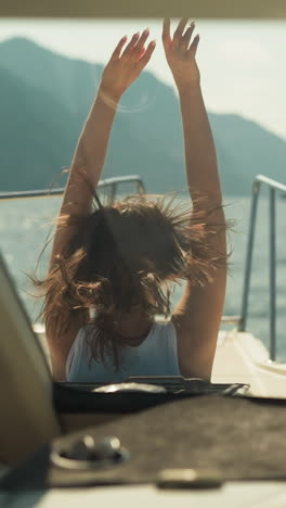 positive female takes moment to stretch arms admiring hills with lush trees from deck of modern motorboat. boat ride on wide sea bay in warm weather