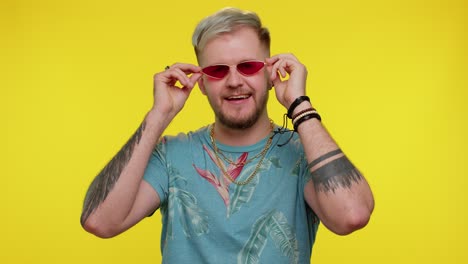 Seductive-cheerful-stylish-man-in-blue-t-shirt-wearing-sunglasses,-charming-smile-on-yellow-wall