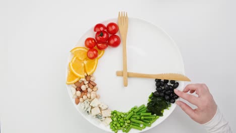 colorful food and cutlery arranged in the form of a clock on a plate. hand takes an olive. intermittent fasting, diet, weight loss, lunch time concept.