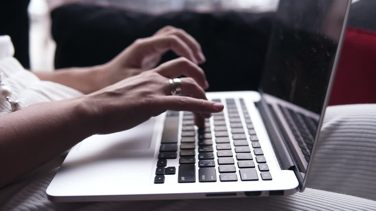 Aim footage of a young women's hands typing on a silver laptop. Computer is on her legs. Black screen