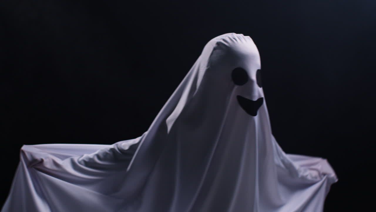 ребёнок, одетый в костюм призрака, делает трюк или угощение на хэллоуин, пугая людей перед черным фоном студии.