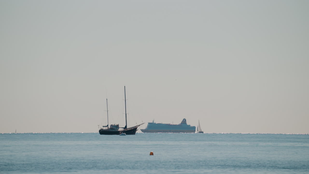 Wide shot of a calm sea with a sailboat and a large cruise ship sailing in the distance under a pale blue sky