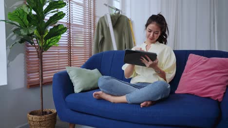 asian attractive female woman wear casual cloth home isolate quarantine enjoy watching online movie on tablet on sofa in living room home interior background home sweet home clam peaceful weekend day