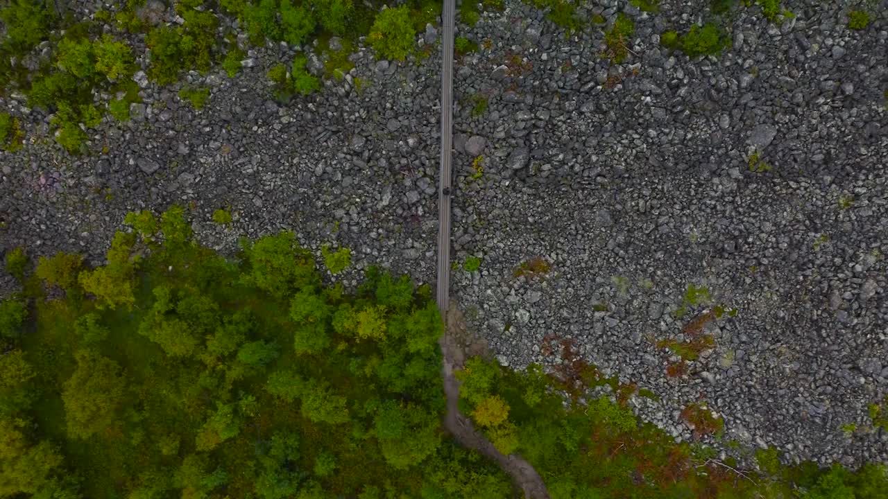 Top down aerial drone footage of a person walking on a wooden hiking trail or a bridge over some rocky and granite gravel covered terrain during cloudy day at Sweden. green forest trees on the bottom.
