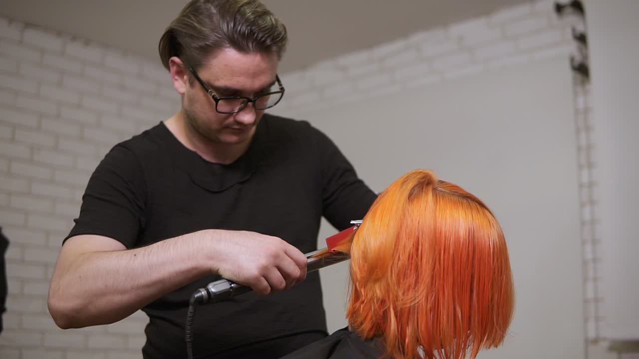 Male professional hairdresser is straightening bright red woman's hair using a hairstraightener in hair salon. Slow Motion shot